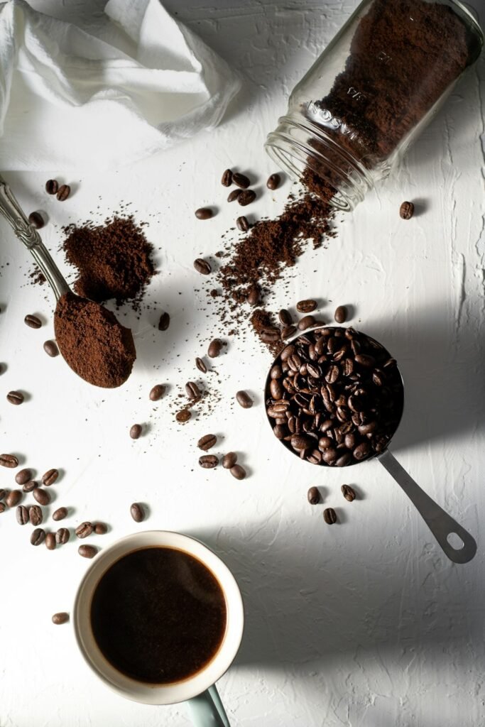 Café moulu dans une cuillère, grains de café et tasse de café noir sur une table blanche